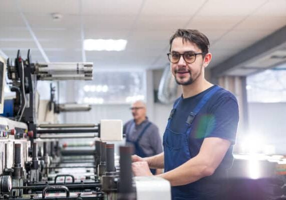 Portrait of content young printing specialist in glasses standing at printing press