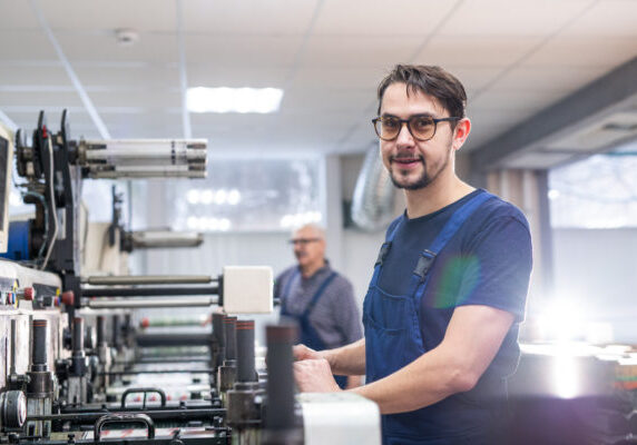 Portrait of content young printing specialist in glasses standing at printing press